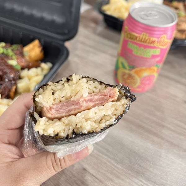 Hand holding a Spam Musubi, with a pink Hawaiian Sun drink and other Hawaiian food in the background.
