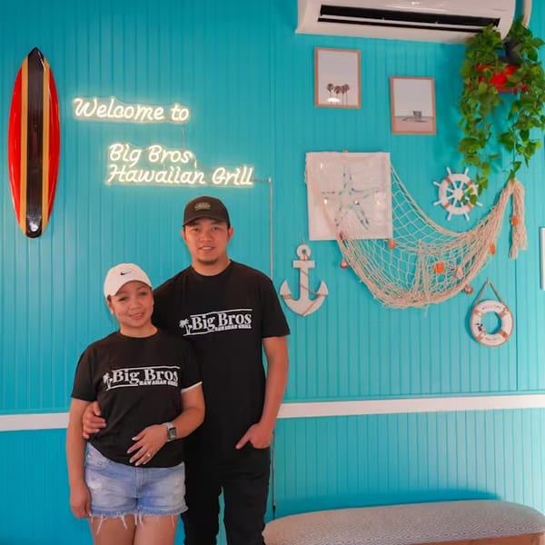 Two Big Bros Hawaiian Grill employees stand in front of a teal wall with a neon sign and nautical decor.