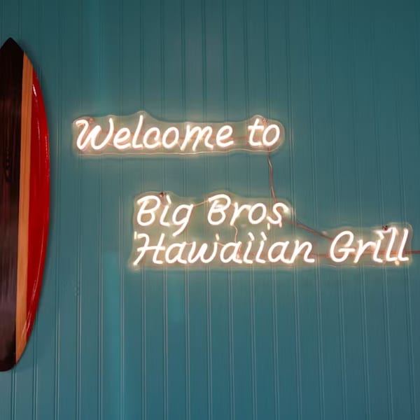 Neon sign on a teal wall reads "Welcome to Big Bros Hawaiian Grill" next to a wooden surfboard.